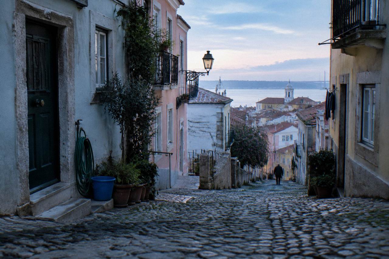 Narrow cobbled street in Alfama with pastel buildings and a glimpse over Lisbon rooftops